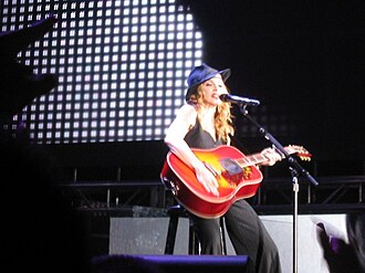 Image of a blonde woman sitting on a stool. She's wearing a black hat, black sleeveless shirt and black pants. She's holding an orange acoustic guitar in her hands. A microphone stands before her. Gray bright imagery can be seen in the background.